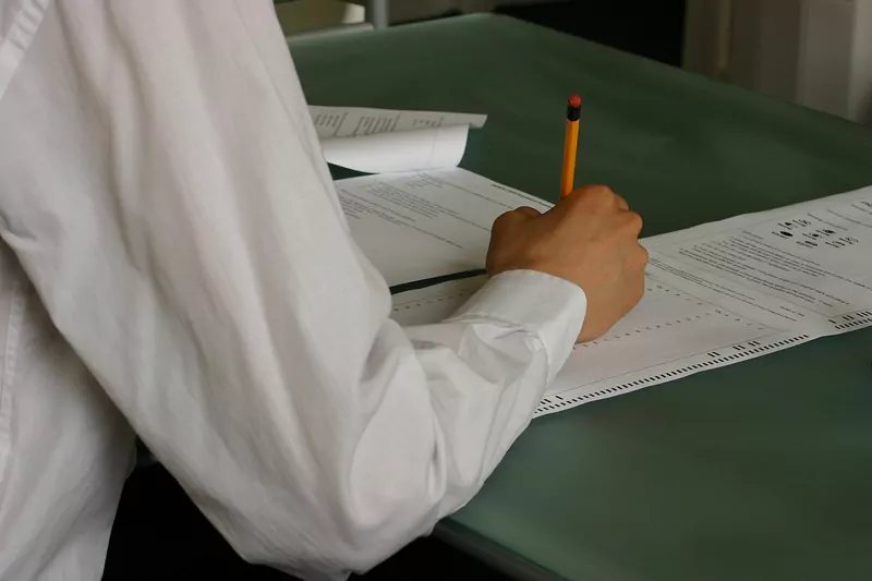 A student in a white long-sleeved shirt writing on an exam paper with a pencil at a green desk