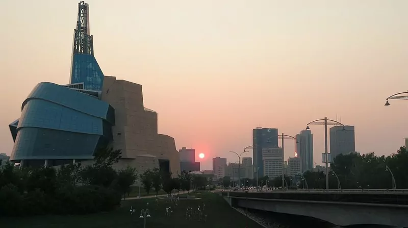 Downtown Winnipeg's towers at sunset, with the glass and stone Canadian Museum for Human Rights on the left and a bridge in the foreground