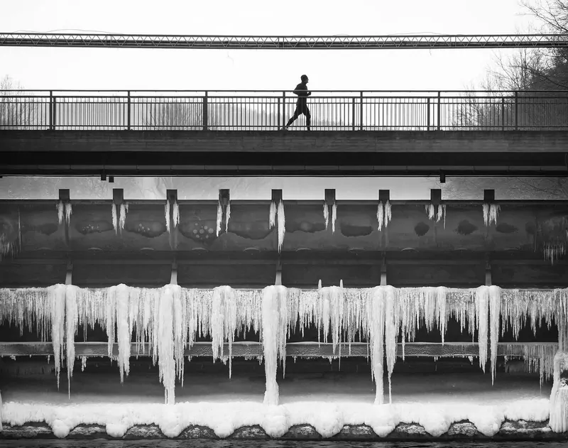 A lone jogger crossing a footbridge in winter, above a weir covered in long icicles