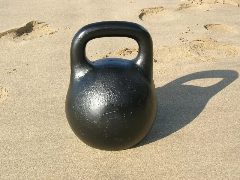 A black cast-iron kettlebell standing on pale sand, with its shadow falling to the right