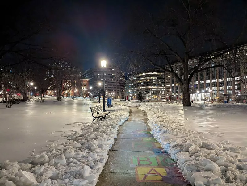 A cleared path through a snowy city park at night in Washington, D.C., with piles of snow on both sides, a bench and building lights beyond