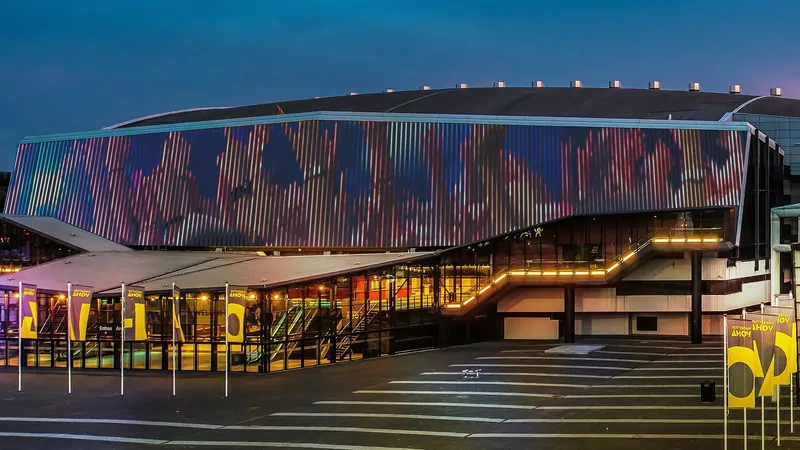 The Rotterdam Ahoy arena at dusk, its LED-lit facade glowing in pink, blue and orange above a plaza with yellow-and-black Ahoy flags