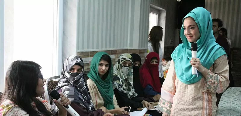 A woman in a turquoise headscarf speaks into a microphone to a group of seated women at a World Mental Health Day session