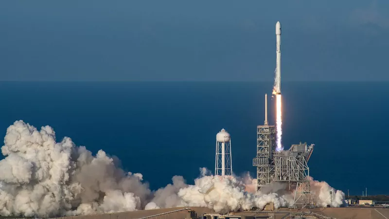 A white Falcon 9 rocket lifting off from a launch tower beside the ocean, with a large cloud of exhaust steam below it