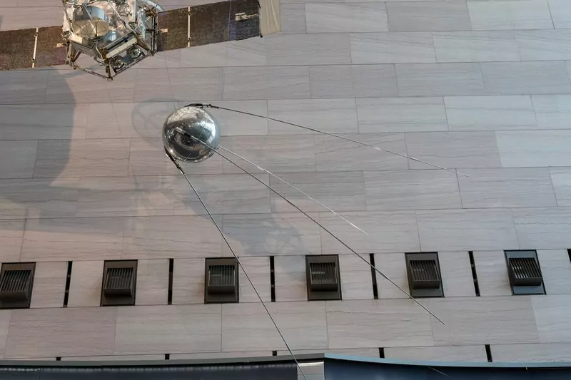 A shiny metal sphere with four long antennas hanging against a stone wall inside a museum